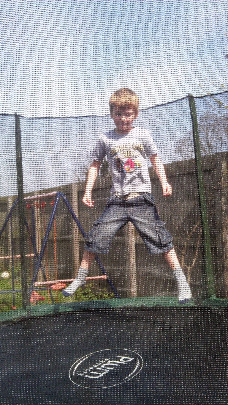 A young boy in a T-shirt and shorts is jumping on a trampoline in a garden for Flashback Friday, with his legs spread apart. A fence and climbing frame are in the background, and netting surrounds the trampoline.