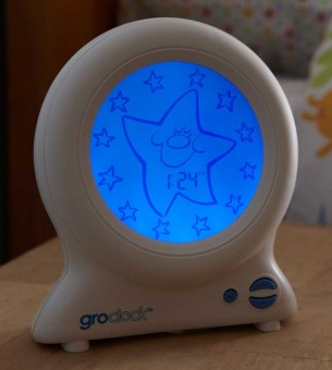 A white Gro-Clock showing a blue-lit screen with a large sleeping star, smaller stars, and a digital time display reading 7:24 sits on a wooden surface—perfect for bedtime routines. Enter now for your chance to win Gro-Clock!.