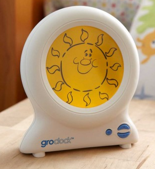 A Gro-clock sleep trainer for children sits on a wooden surface, displaying a smiling sun with rays on its yellow screen. The white device has blue buttons and the Groclock logo—enter now for a chance to win Gro-clock!.