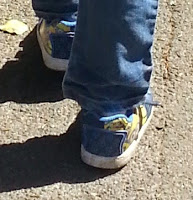 A person with summer style, wearing blue jeans and blue trainers with yellow cartoon designs from Shoe Zone, walks on a sunlit, textured pavement.