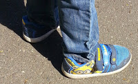 A child wearing blue jeans and blue Minions trainers from Shoe Zone stands on a sunlit pavement, stepping out in style this summer.