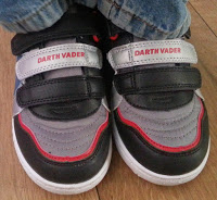 A close-up of a child showing off their summer style in black, grey, and red Darth Vader trainers from Shoe Zone, standing on a wooden floor and wearing blue jeans.