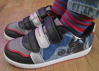 A child showcases summer style in blue jeans and colourful striped socks, sporting black, grey, and red Velcro Star Wars trainers from Shoe Zone whilst standing on a wooden floor.