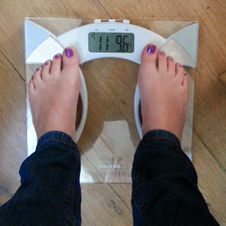 Person with purple painted toenails standing on a glass digital bathroom scale showing a weight of 11 stone and 9.6 pounds, possibly tracking progress for the SlimFastChallenge, on a wooden floor.