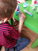 A young child in a red and black striped shirt kneels on the floor, crafting at a green table with felt-tip pens and glue sticks scattered on top, making Half Term fun and creative.
