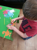 A child in a red shirt enjoys crafting at a small green table, drawing on an orange paper dinosaur amid other colourful cut-outs and pencils—perfect fun for half-term.