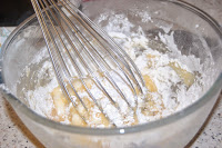 A metal whisk mixing flour and wet ingredients in a clear glass bowl, sitting on a speckled worktop—the first step in tackling the Challenge inspired by Heston’s iconic Pear Tarte Tatin.