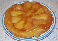 A golden brown Pear Tarte Tatin with caramelised pear halves arranged on top, inspired by a Heston challenge, sits on a white plate with blue stripes on a speckled worktop.