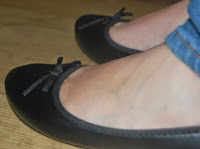 A close-up of a person's feet in black ballet pumps with small bows, paired with blue jeans, standing on a wooden floor—perfect summer style from Shoe Zone.