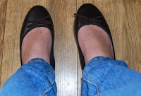 A person showcases summer style in blue jeans and black ballet pumps with bows from Shoe Zone, standing on a wooden floor.