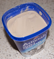 A blue plastic tub of Astonish oven and cookware cleaner, with the lid off, reveals smooth, pale cream cleaning paste inside—perfect for Spring Cleaning. The tub sits on a speckled worktop.