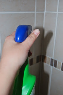 A hand holding an Astonish green spray bottle with a blue trigger, positioned in front of tiled bathroom walls—perfect for your spring cleaning routine.