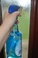 A hand holds an Astonish spray bottle filled with blue cleaning liquid, pointed at a glass door or window—ready for some Spring Cleaning.