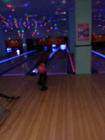 A person is bowling in a dimly lit alley with colourful lights on the ceiling and neon lights highlighting the lanes during James's 6th birthday celebration, about to release a pink bowling ball towards the pins.