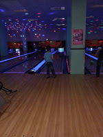 A person in a blue shirt prepares to bowl at a glowing bowling alley during James's birthday celebration. Colourful reflections shine on the ceiling, and several lanes and another bowler are visible in the background.