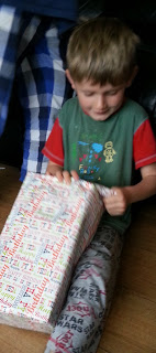 On this Silent Sunday, a young boy sits on the floor, smiling as he unwraps a large, colourful gift box. He wears pyjamas and a green shirt with red sleeves, with a blue and white chequered blanket in the background.