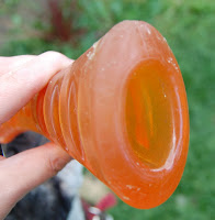 A hand holds an orange, translucent, cone-shaped Bionic Dog Toy with a wide, rounded opening, outdoors with green grass in the background.