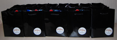 A row of glossy black gift bags with white circular labels on the front, neatly lined up on a light-coloured surface—perfect party favours for James's sixth birthday, each bag filled with surprises for guests celebrating him turning 6.