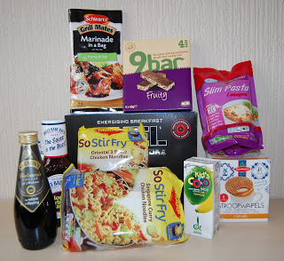 A selection of packaged food products from a recent Degustabox, including noodles, marinade, syrup, snack bars, pasta, tea, caramel waffles, and a children's chocolate drink—perfect for a subscription box discovery—arranged on a light surface against a textured wall.