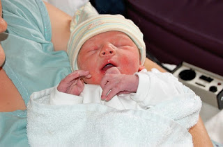A newborn baby wearing a striped hat and wrapped in a white blanket is being held by an adult, possibly in a hospital setting. It’s a touching flashback Friday moment with the baby’s eyes closed and mouth slightly open.