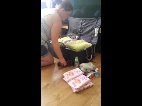 A woman kneels on the floor packing a suitcase filled with clothes, ready for the Great Pack Off. Baby wipes, toys, baby lotion, and toiletries from Holiday Extras are neatly arranged on the wooden floor in front of her.