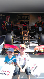 Two young boys in racing caps and shirts sit in front of a black and gold Lotus F1 car display, holding Nissan flags. Large images of racing drivers are visible on the backdrop—a true Flashback Friday moment for motorsport fans.