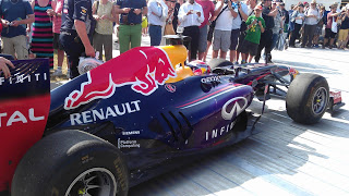 Flashback Friday: A Red Bull Racing Formula 1 car with Renault and Infiniti logos is on display outdoors, surrounded by a crowd of people taking photos and observing.