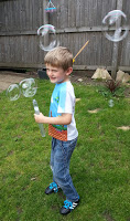 A young boy in a blue and white t-shirt and jeans stands on grass, smiling as he holds a bubble wand, enjoying sunshine. Several large bubbles float in the air around him. A wooden fence is in the background.