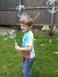 A young boy stands on grass blowing bubbles on a Silent Sunday, holding a bubble wand and container. He smiles mischievously in jeans and a colourful t-shirt as bubbles float around him, with a wooden fence in the background.