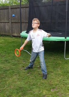 A boy in glasses stands on grass, holding an orange racquet and preparing to hit a blue ball. He is making the most of the sunshine with outdoor activities near a trampoline and a wooden fence.