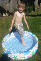 A young boy stands in a small inflatable paddling pool filled with water in a sunny garden, making the most of the sunshine in patterned swimming trunks and smiling at the camera.