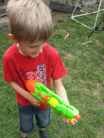 Young boy in a red shirt and shorts holding a bright green and orange water pistol, standing on grass in a garden, making the most of the sunshine with fun outdoor activities.