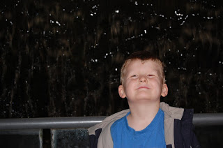 A young boy with light skin and short hair, wearing a blue shirt and jacket, stands in front of a dark, watery background, smiling slightly with his eyes closed—a perfect flashback Friday moment.