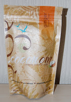 A tan and orange resealable pouch labelled "Coconut Ginger" stands upright against a light-coloured background, featuring a decorative floral design and small blue birds—perfect for your next food subscription or snack subscription box discovery.