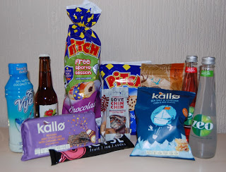 A variety of snacks and drinks from a snack subscription box, including crisps, crackers, chocolate, coconut water, bottled drinks, and snack bars, are arranged on a light-coloured surface in front of a beige wall.