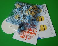 A small pile of puzzle pieces in a blue net, two circular pieces, and a Minion-themed instruction sheet for a 3D Minion Puzzle on a green background.