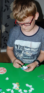 A boy wearing glasses and a grey shirt is focused on assembling a Ravensburger Puzzle.