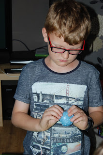 A young boy with glasses looks down, focusing on a small blue 3D Minion Puzzle in his hands. He is wearing a T-shirt with an image of the Golden Gate Bridge and the word "San Francisco" on it.