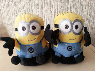 Two plush toys resembling Minions, with yellow bodies, blue dungarees, black gloves, and big round eyes, sit side by side on a light-coloured surface against a textured wall, looking just like adorable Minion slippers or Stompeez Slippers.