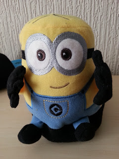 A plush toy resembling a Minion character from "Despicable Me," with large embroidered eyes, blue dungarees, and black gloves, sits on a light-coloured surface—perfect for pairing with cosy Minion slippers or Stompeez slippers.