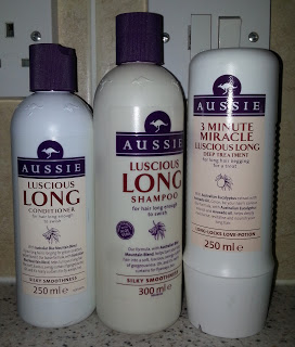 Three bottles of Aussie hair care products—Luscious Long Conditioner, Shampoo, and 3 Minute Miracle Deep Treatment—are arranged on a countertop against a tiled wall, available now at Chemist Direct.