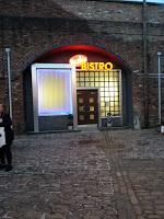 A neon sign reading "BISTRO" above a doorway set in a brick building on the Coronation Street Tour, with a glowing window on the left and cobbled pavement in front; two people stand partially visible on the left side.