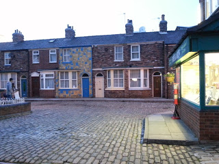 A cobblestone street lined with brick terraced houses featuring colourful doors and windows, reminiscent of those seen on a Coronation Street Tour, with a shop displaying goods on the right side of the image.