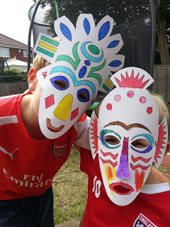 Two children wearing red football shirts and colourful, homemade paper masks pose together outdoors on a grassy lawn for Silent Sunday. Trees, houses, and a trampoline are visible in the background.