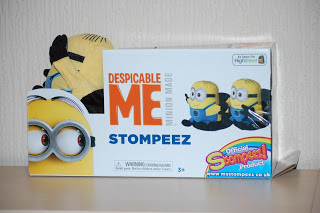 A box of Despicable Me Minion Stompeez Slippers, featuring fun Minion images and colourful branding, sits on a white shelf with a partial plush Minion toy visible behind the box.