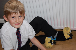 Smiling young boy in a white shirt and striped tie sits on the floor next to someone wearing black trousers and fun yellow Minion Stompeez slippers, with a white radiator in the background.