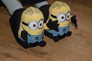A pair of feet wearing Minion Stompeez slippers, shaped like the yellow characters from the "Despicable Me" films, rests on a wooden floor.