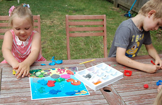 Two young children sit at a wooden table outside, playing with colourful Plasticine Softeez and shaped moulds. A board with sea creatures and an open tray of clay are on the table. Grass and a fence complete the playful scene.