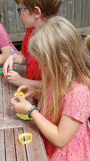 A young girl with long blonde hair and a pink dress plays with yellow Plasticine Softeez at a wooden table next to a boy in a red shirt and spectacles.