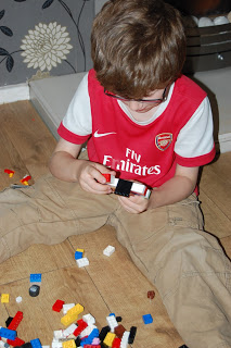 A young boy wearing glasses and an Arsenal football shirt sits on a wooden floor, focused on building his Dream Lego House with colourful LEGO bricks scattered around him.